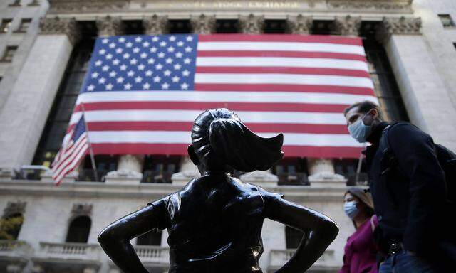 Pedestrians walk by the Fearless Girl Statue and a giant American Flag that hangs outside at the New York Stock Exchange