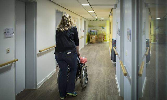 Eine Betreuerin schiebt eine aeltere Frau im Rollstuhl durch einen Gang in einer Pflegeeinrichtung, aufgenommen in Berl