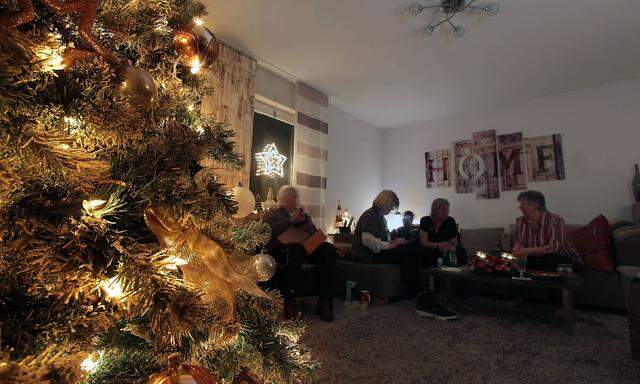Heiligabend: beleuchteter Weihnachtsbaum steht in einem Wohnzimmer mit Fanilie im Hintergrund gemuetlich auf dem Sofa si