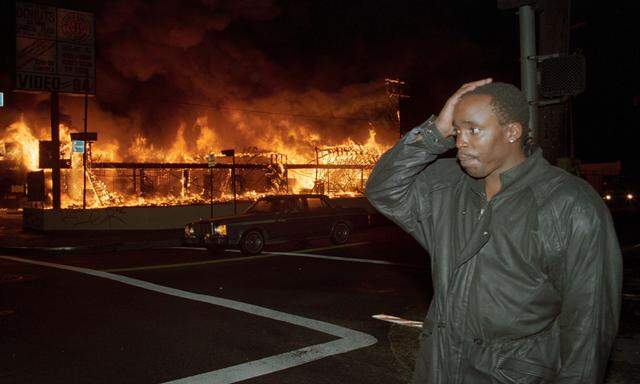 A businessman holds his hand to his head as the store he owns burns down across the street from him during the Los Angeles Riots