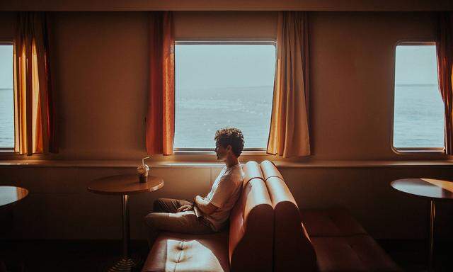 Side view thoughtful young male wearing casual summer clothes sitting on soft couch while travelling in ferry on ripplin
