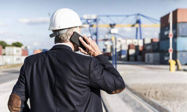 Businessman at cargo harbour wearing safety helmet using smartphone model released Symbolfoto PUBL