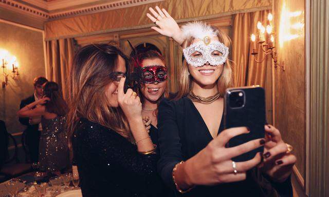 IMAGO Creative: Guten Rutsch  Glad female friends in masquerade masks taking self portrait on smartphone while having di