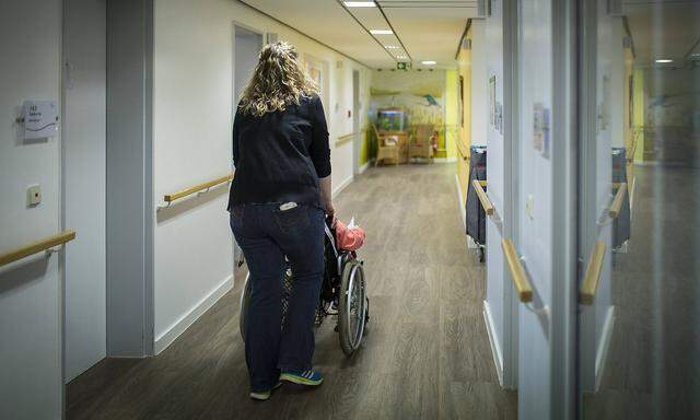 Eine Betreuerin schiebt eine aeltere Frau im Rollstuhl durch einen Gang in einer Pflegeeinrichtung