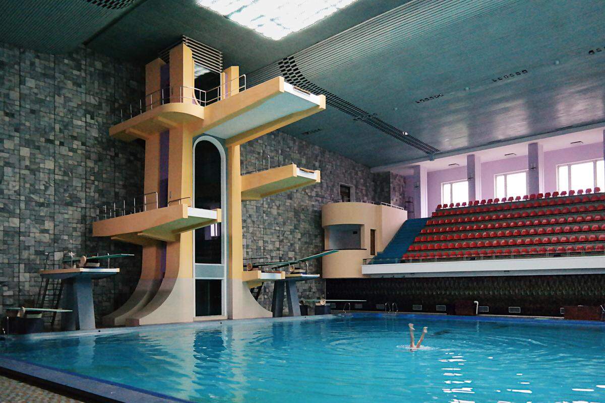 Auf den Sprungturm im Schwimmbad gelangt man mit einem futuristisch anmutenden mechanischen Aufzug, der sich in einem mit Rauchglas verkleideten Schacht befindet.