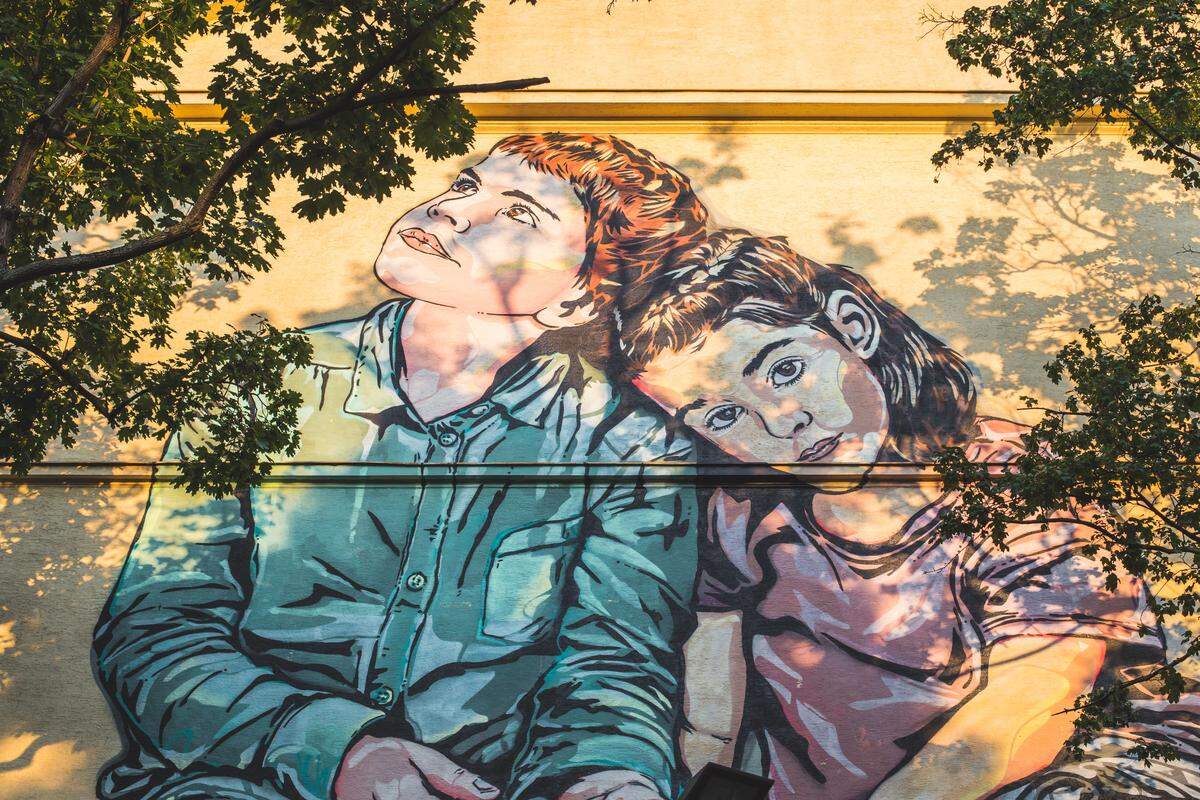 ...Ähnliches symbolisiert auch das Mural des Künstlerduos Jana und Js in der Währingerstraße. Als Anlehnung an die Bezirksvorsteherin mit iranischen Wurzeln, zeigt das Mural ein Kinderpaar mit Migrationshintergrund. Die Kinder stehen für die perfekte Zukunft, in der weder Klassen-, noch Gender- oder Rassenunterschiede gemacht werden sollen. Die Rose in den Händen der beiden symbolisiert Entfaltung. 