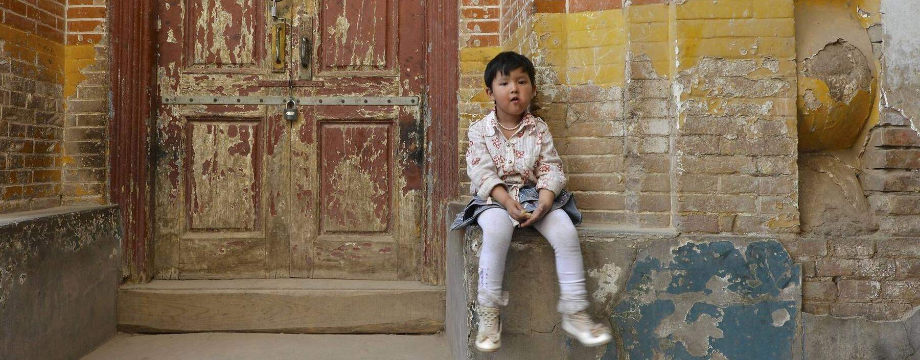 Kleines Mädchen sitz auf Backsteinfassade in den Gassen eines uigurischen Wohnviertels,