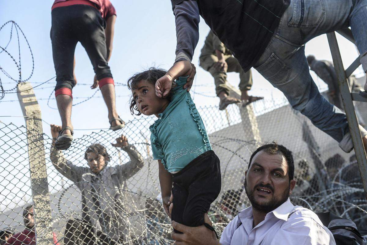 Bulent Kilic, Türkei, 2015, für Agence France-Presse Ein Flüchtling hält ein Mädchen an der türkischen Grenze bei Sanliurfa, am 14. Juni 2015.  Bei Ausbruch des Syrien-Konflikts (2011) befolgte die Türkei erst eine Politik der offenen Tür für Menschen, die vor der Gewalt flohen. Im Juni 2015 führten neue Kämpfe in Nord-Syrien zu einem Anstieg der Grenzübertritte, als die Zivilbevölkerung vor den Luft- und Bodenkämpfen floh. Nach UNHCR-Angaben haben die Kämpfe dazu geführt, dass in der ersten Julihälfte über 23.000 Flüchtlinge, zu 70 Prozent Frauen und Kinder, in die Türkei flohen. Als dieTürkei Maßnahmen zur Eindämmung des Flüchtlingszustroms ergriff, suchten Menschenandere Einreisewege.