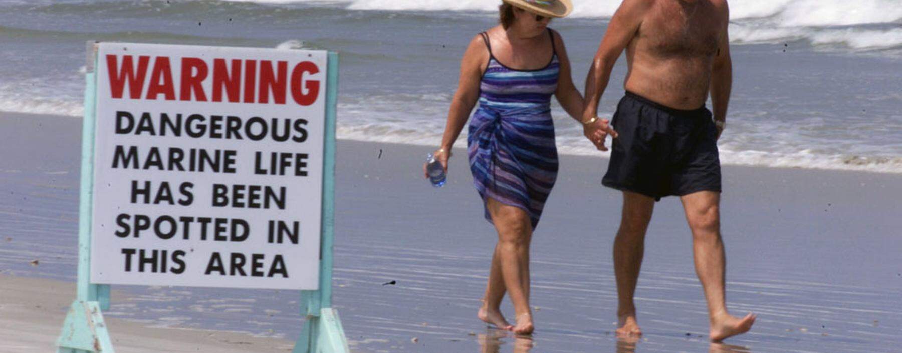 BEACHGOERS WALK NEAR A SIGN OF DANGEROUS MARINE LIFE.