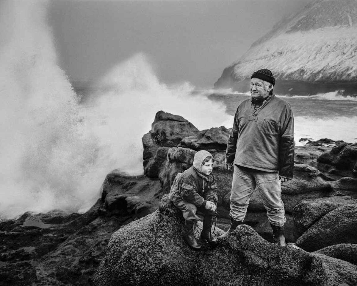 Poul Sigur Cristiansen steht mit seinem Enkelkind in Regenkleidung draußen am Meer. Gjóvgv, Insel Eysturoy, Färöer.