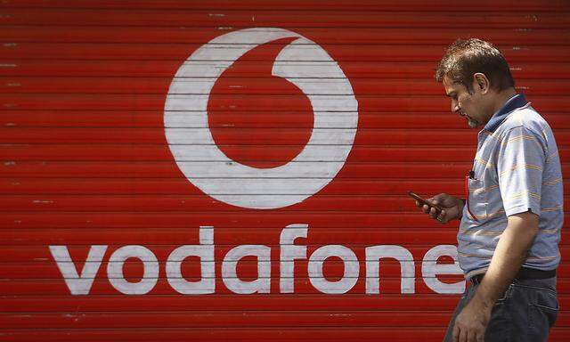 A man checks his mobile phone as he walks past a shop displaying the Vodafone logo on its shutter in Mumbai