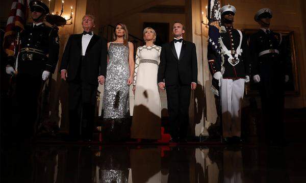 Trump und Macron dinierten im schwarzen Smoking, die Ehefrauen erschienen in langen Roben. Melania Trump trug ein silberfarbenes Kleid von Chanel, Brigitte Macrons ein cremefarbenes Gewand mit goldfarbenem Besatz von Louis Vuitton.