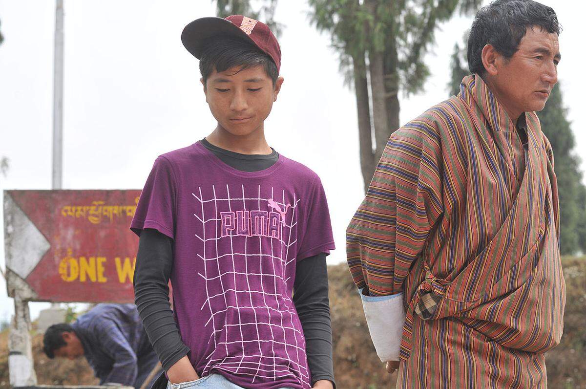 Ein junger Bhutanese im Markenshirt steht am Markt in Tsirang neben einem erwachsenen Mann im Gho. In ihrer Freizeit ziehen viele Jugendliche lieber „westliche“ Kleidung an, die sie im Fernsehen sehen.