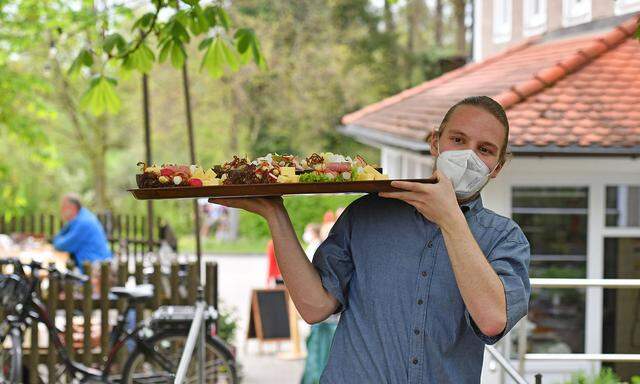 Ein Kellner traegt ein Tablett mit bayerischen Schmankerl,Speisen zu den Tischen-er traegt einen ffp2 Mundschutz,Maske.