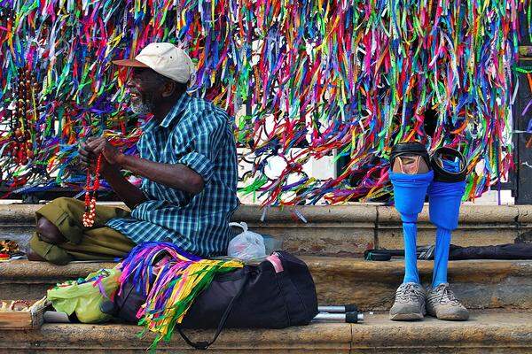 Carlos Henrique Reinesch, Brasilien: "A Blast of Colors"  Ein Mann ohne Beine lächelt, während er Armbänder verkauft. Hinter ihm ist eine Kirche, der Zaun voller Bändchen. Sie gehören zu einer lokalen Tradition: Wer ein Band anbringt, darf sich etwas wünschen.
