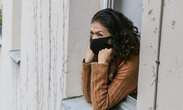 frau in quarantaene mit mundschutz schaut depressiv aus dem fenster ihrer wohnung *** woman in quarantine with mouth gua