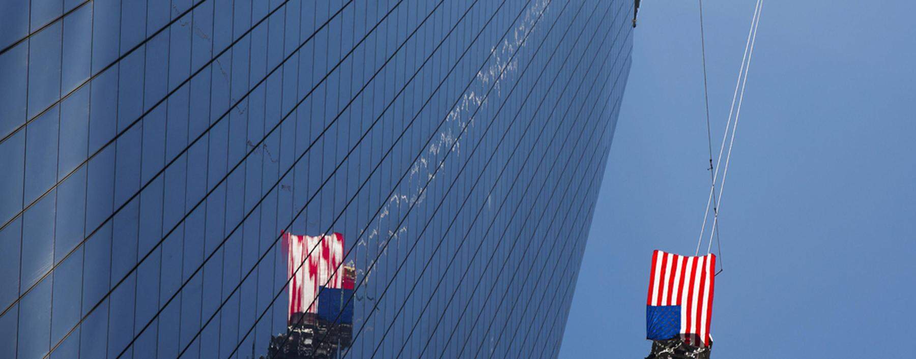 A U.S. flag flutters in the wind as the final piece of a spire is lifted to the top of One World Trade Center in New York