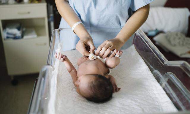 Mother wearing hospital gown changing newborn baby diaper while standing in hospital model released Symbolfoto MPPF01480