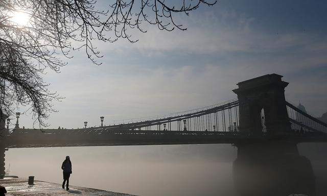 Woman walks front of the Chain Bridge which is wrapped in a veil of fog as the sun rises above the Danube river in Budapest