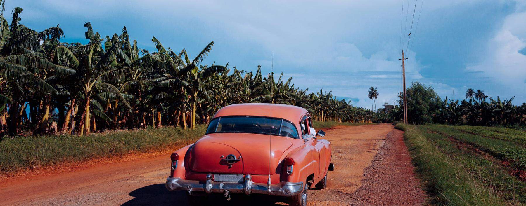 Sandy dirt red road with old vintage car with green plants on sides and grey cloudy sky on background in Cuba Copyright: