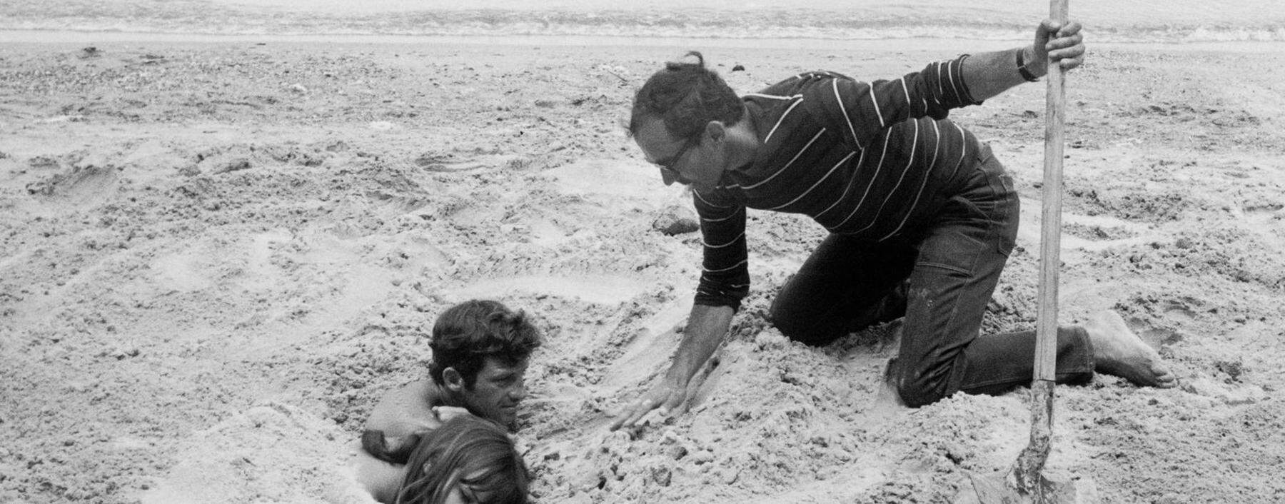 Jean-Luc Godard, wie er bei den Dreharbeiten von „Pierrot le Fou“ („Elf Uhr nachts“, erschienen 1965) seine Darsteller Anna Karina und Jean-Paul Belmondo eingräbt.