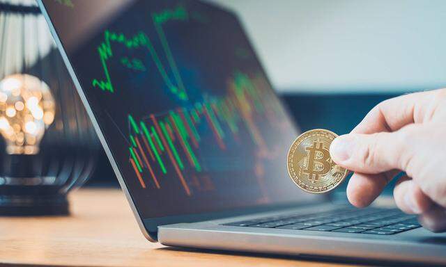 17 June 2022: Hand holding a Bitcoin gold coin BTC cryptocurrency in front of a laptop computer, online trading on stoc