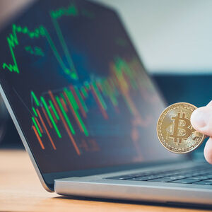17 June 2022: Hand holding a Bitcoin gold coin BTC cryptocurrency in front of a laptop computer, online trading on stoc