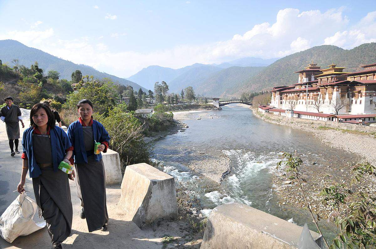 Zwei Schulmädchen in blau-grauer Kira (dem traditionellen bhutanesischen Kleid, das auch als Schuluniform getragen wird) gehen von der Schule nach Hause. Bhutan investiert viel in die Bildung seiner Jugend, die als „Zukunft“ des Landes gilt. Im Hintergrund ist der Punakha-Dzong, einer der berühmtesten Dzongs des Landes, zu sehen.