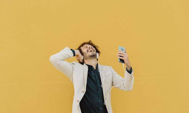 Businessman singing and dancing in front of yellow wall listening music with headphones and smartpho