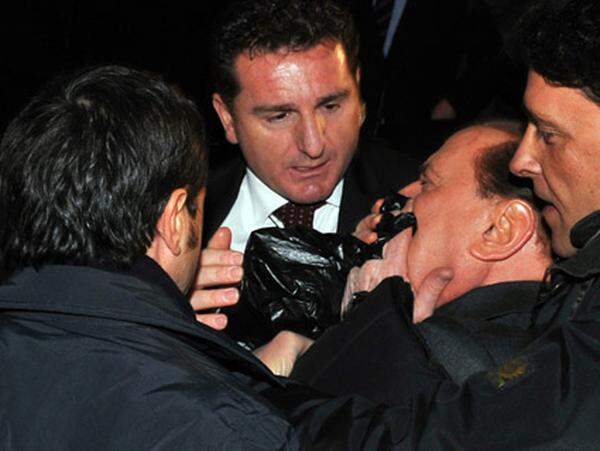 Berlusconi stürzt nach dem Angriff zu Boden. Augenblicke später zeigt er sich blutend den umstehenden Menschen und versichert, dass er nicht schwer verletzt sei.