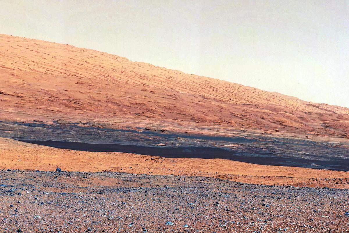 Unweit des dreiachsigen Mars-Rovers fällt die kiesige Ebene in eine Senke ab, der rostrote Streifen in der Mitte ist der Rand eines kleinen Einschlagkraters.